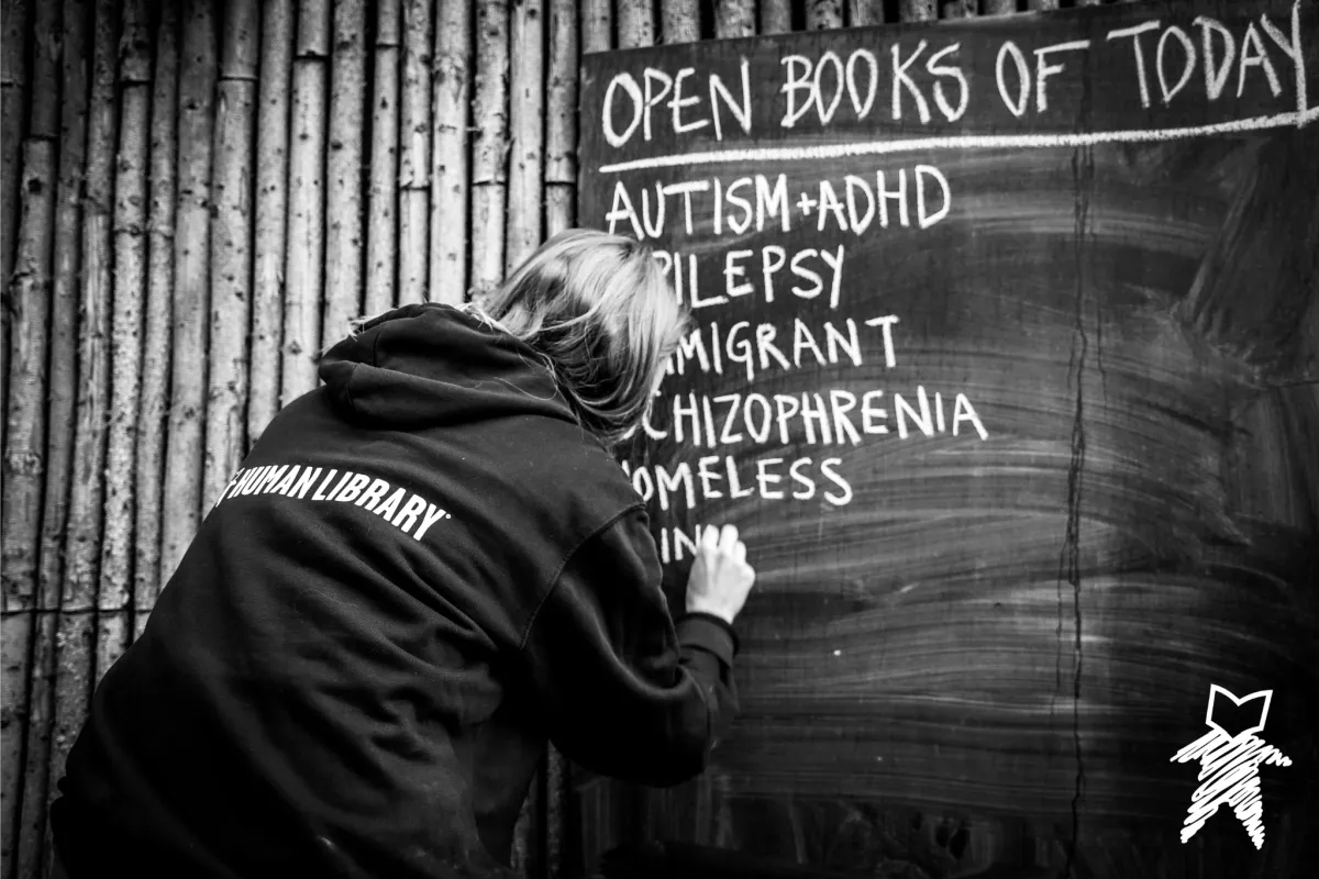 Eine Frau mit einer Kapuzenjacke, auf deren Rücken HUMAN LIBRARY steht, schreibt etwas auf eine Schiefertafel.